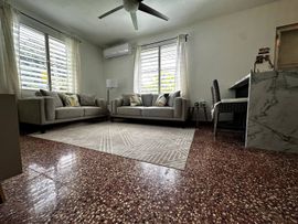 A living room with gray sofas, a patterned rug, a ceiling fan, and speckled flooring near a kitchen counter.