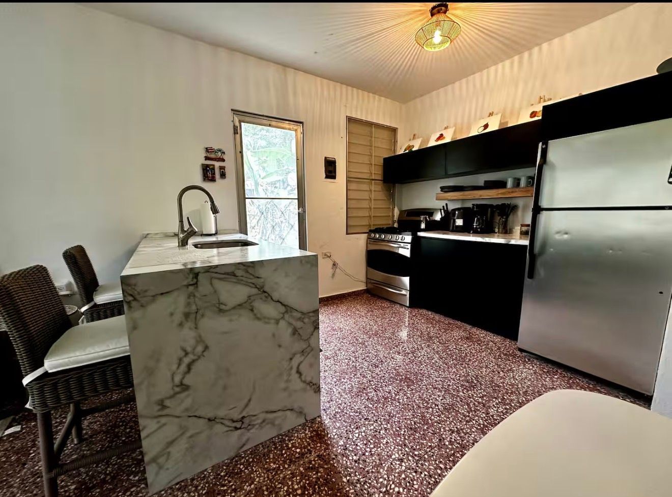 A modern kitchen with a marble-topped island, dark cabinets, stainless steel appliances, and a red terrazzo floor.