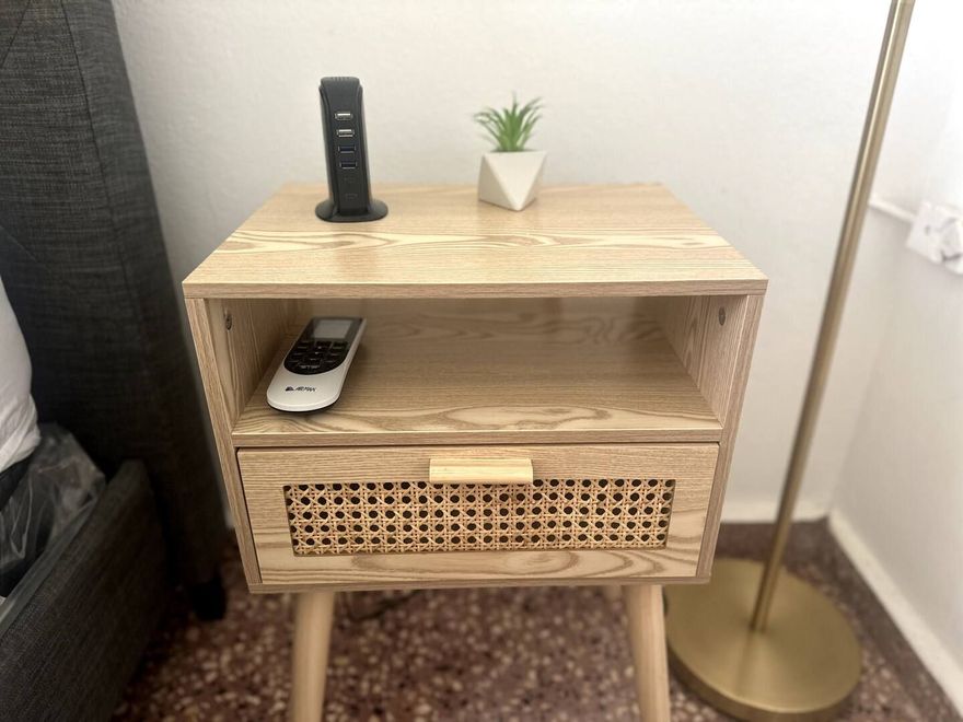 A wooden bedside table with a small open shelf, a rattan drawer, a remote control, a small plant, and a tall lamp.