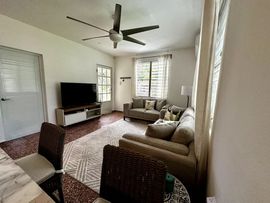 A living room with a beige sofa, television on a white stand, patterned area rug, ceiling fan, and wicker chairs.