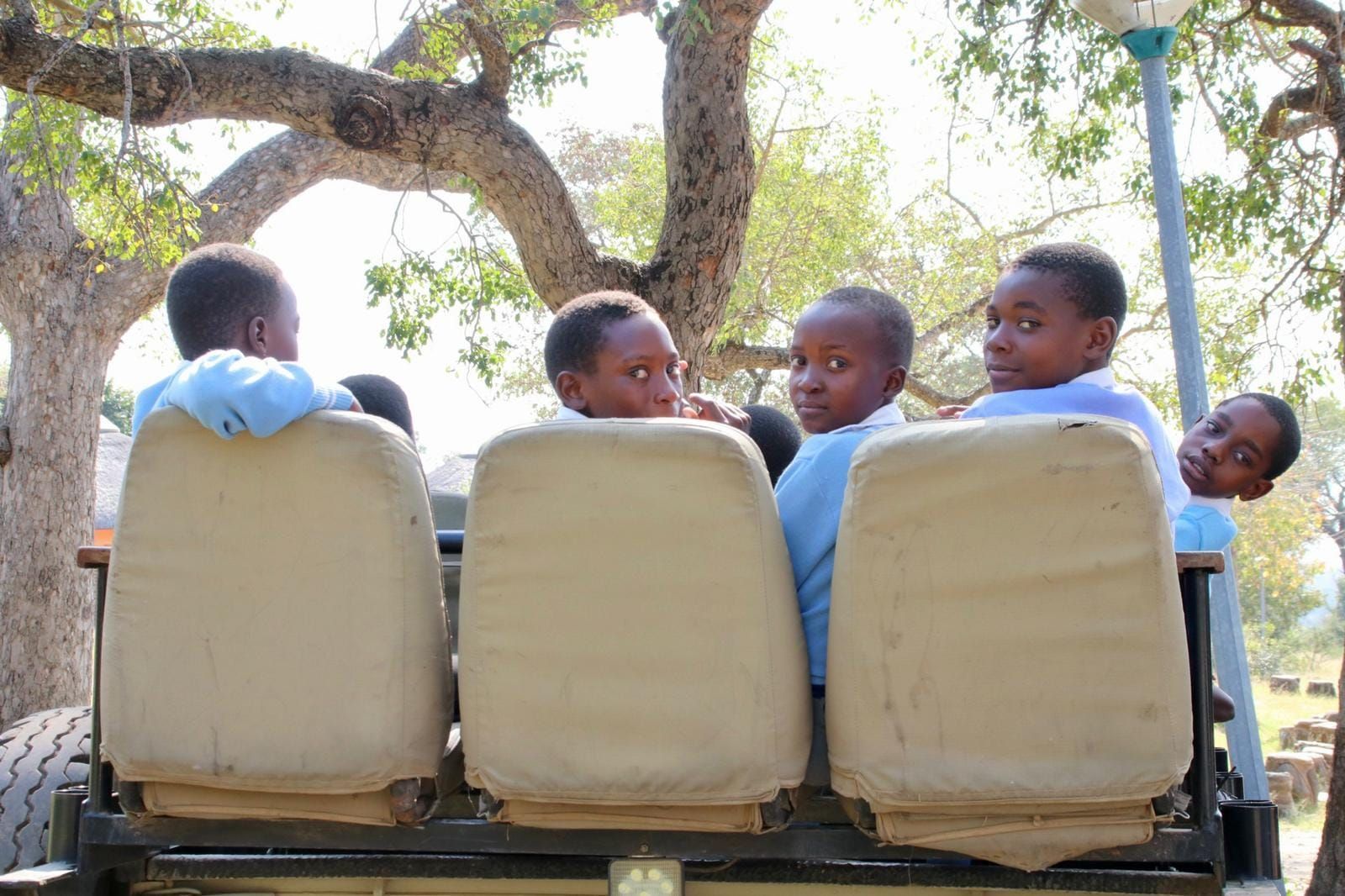 Several people in light blue shirts sit in the back of an open-air vehicle, looking back toward the camera outdoors.