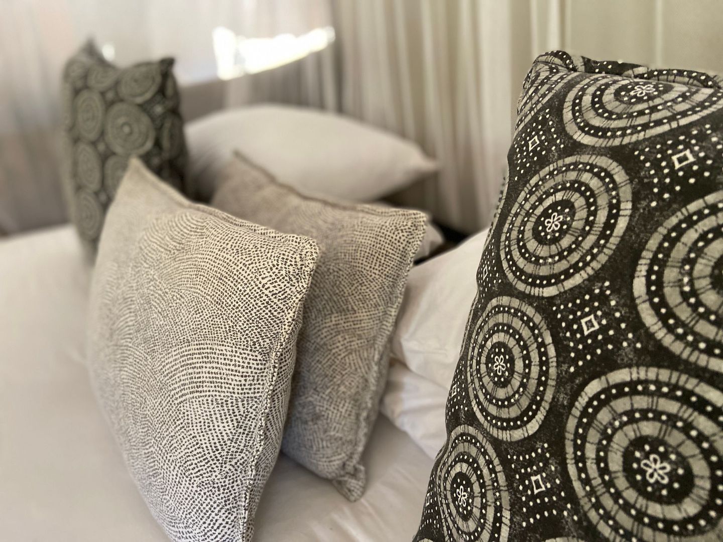 Several decorative throw pillows with varied gray and white patterns rest against a white bedhead in a bedroom.