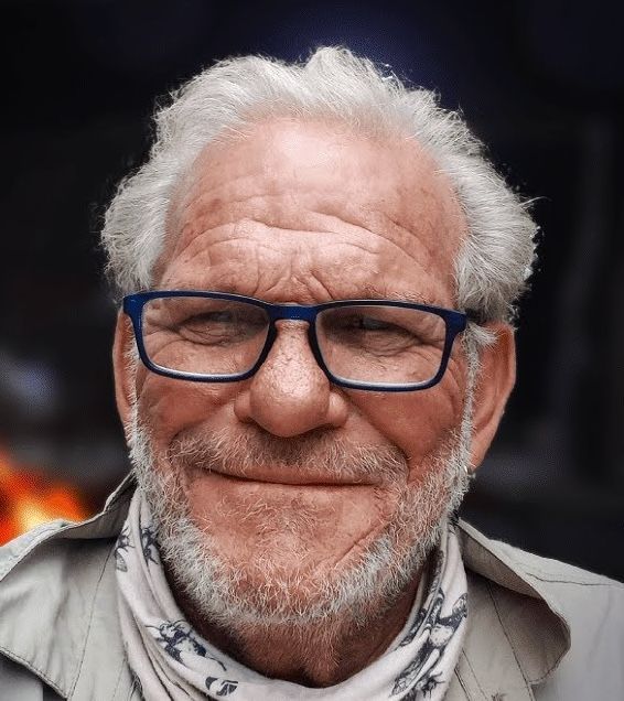 A close-up portrait of a person with white hair and blue-rimmed glasses smiling, wearing a light-colored scarf.