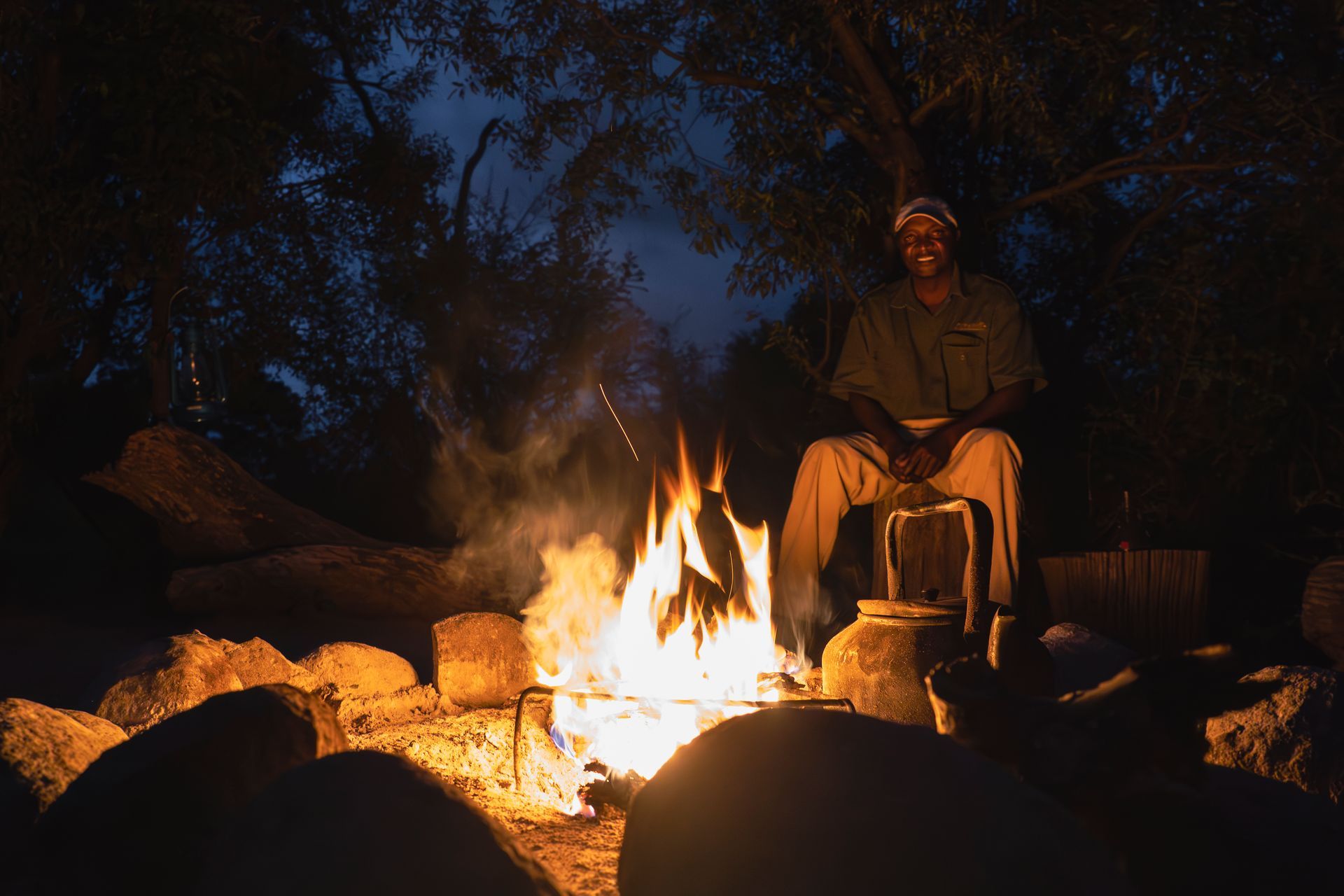 A person sits on a log by a glowing campfire at night, with a kettle resting nearby among rocks in a forest setting.
