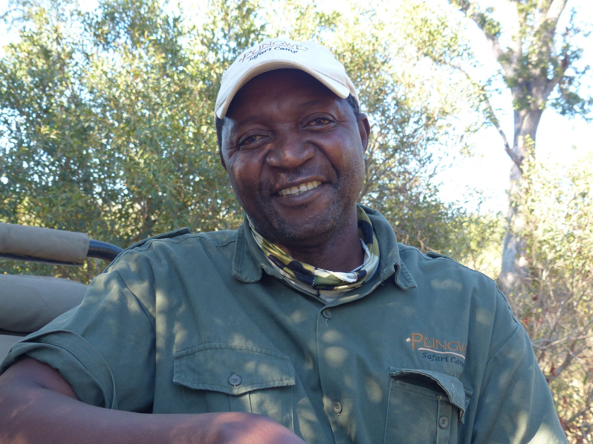 A smiling person in a green uniform and light-colored cap, seated outdoors in a wooded area.