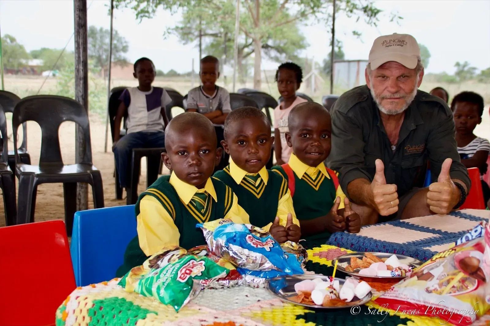 A smiling person gives a thumbs-up while sitting with children in school uniforms at a table set with snacks and cake.