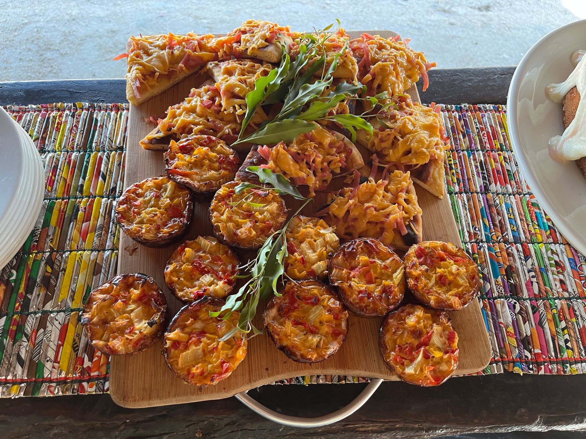 A rectangular wooden board holds a collection of golden-brown appetizers, garnished with green leaves on a patterned mat.