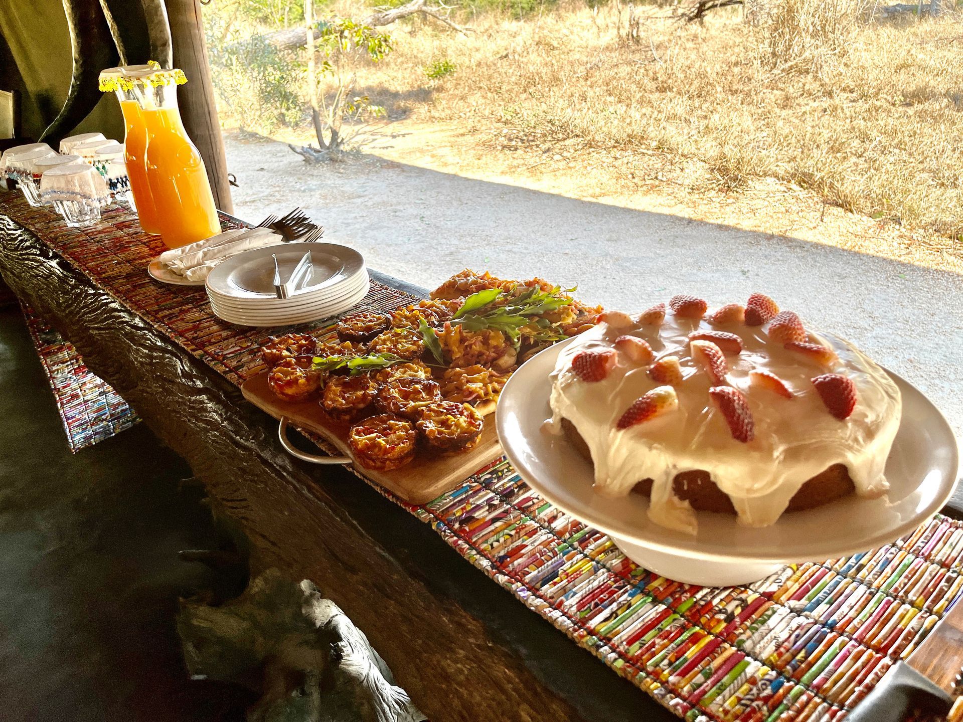 A buffet table displays orange juice, a tray of small pastries, and a strawberry frosted cake overlooking a dry landscape.