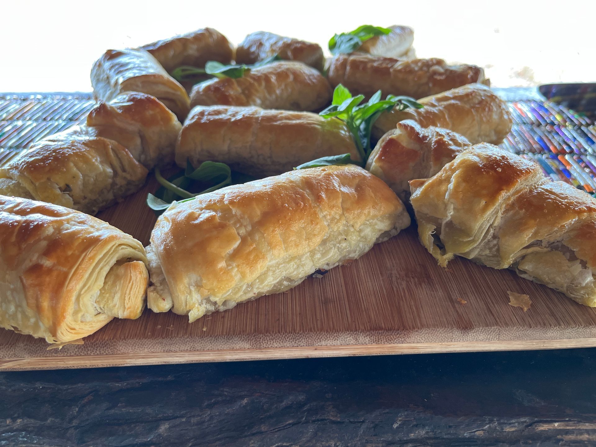 A platter of golden-brown sausage rolls garnished with fresh green leaves on a wooden serving board.