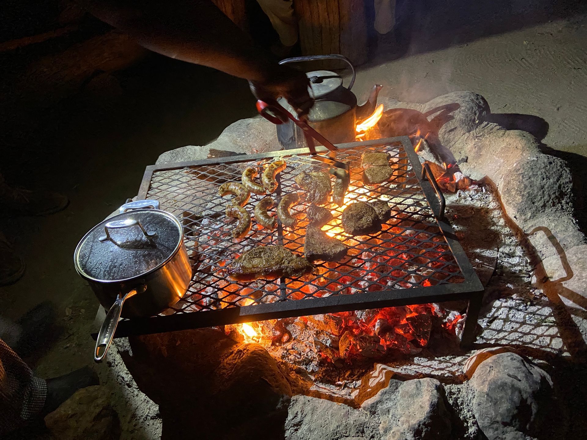 A person uses tongs to grill meat over an open campfire, with a pot sitting on the edge of the grate.