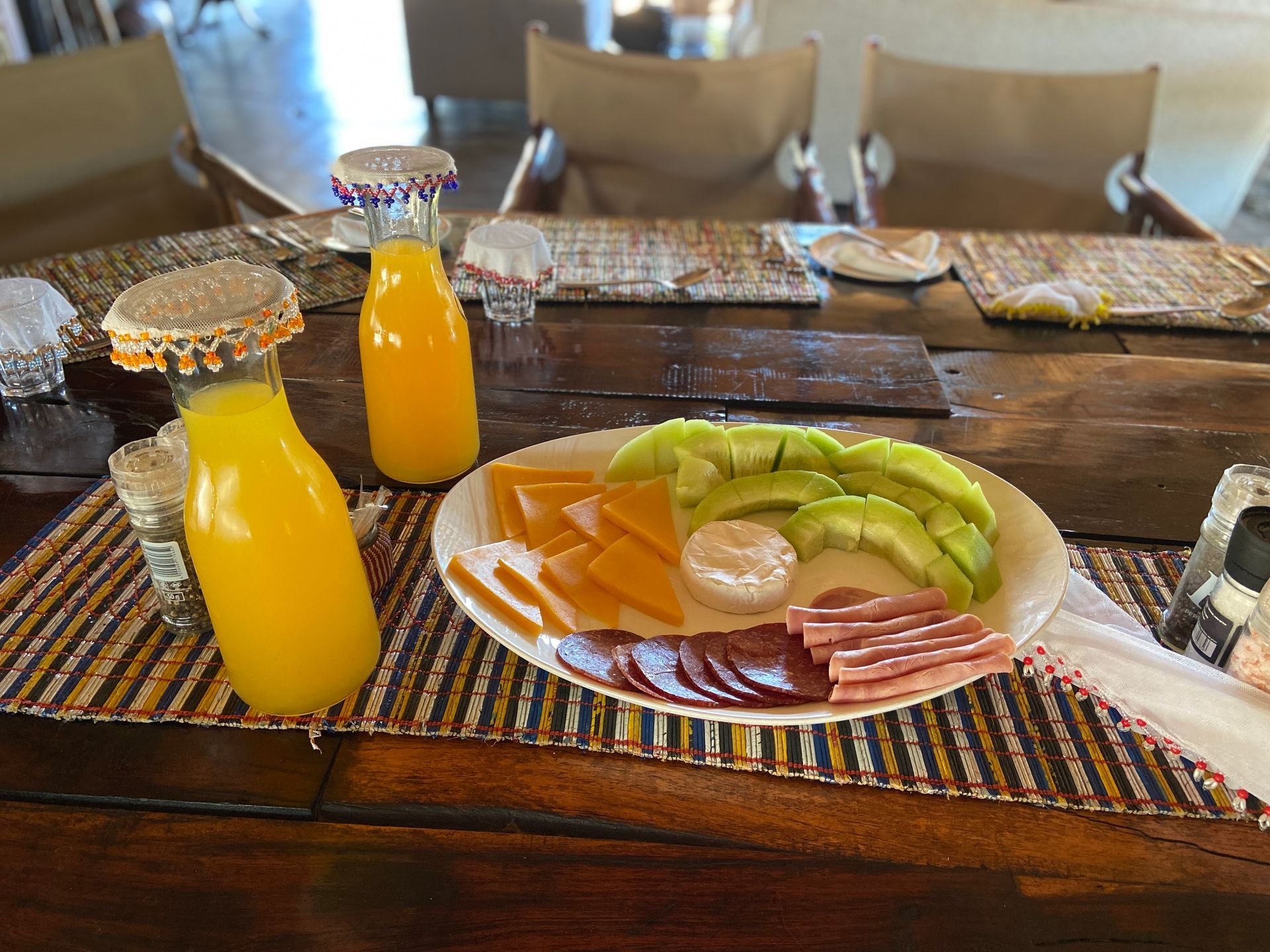 A breakfast table setting featuring two glasses of orange juice and a plate with sliced cheese, avocado, meat, and cheese.