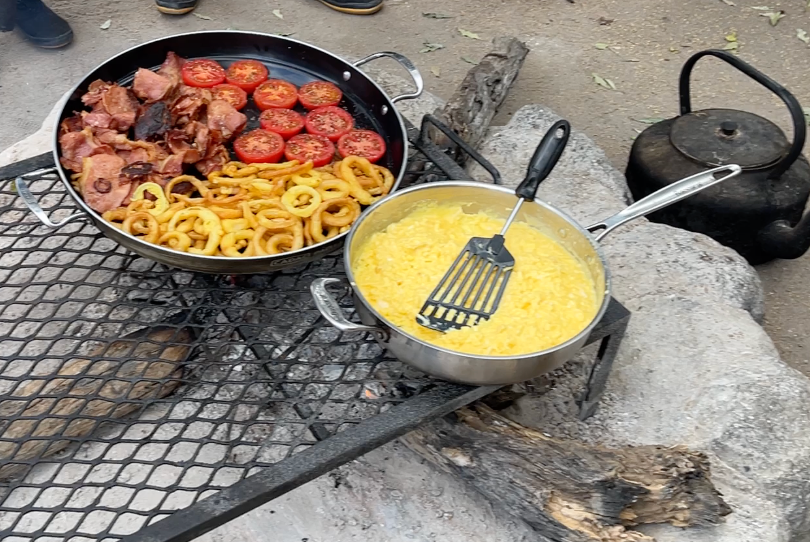 Two pans cooking over an outdoor fire: one with bacon, tomatoes, and onion rings, the other with yellow scrambled eggs.
