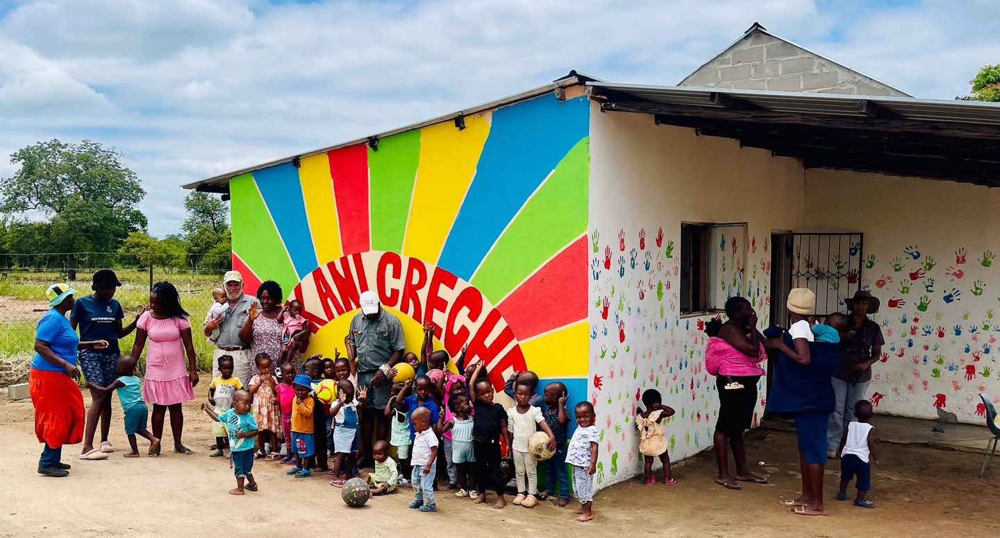 A group of children and adults standing in front of a building with a colorful sunburst mural labeled
