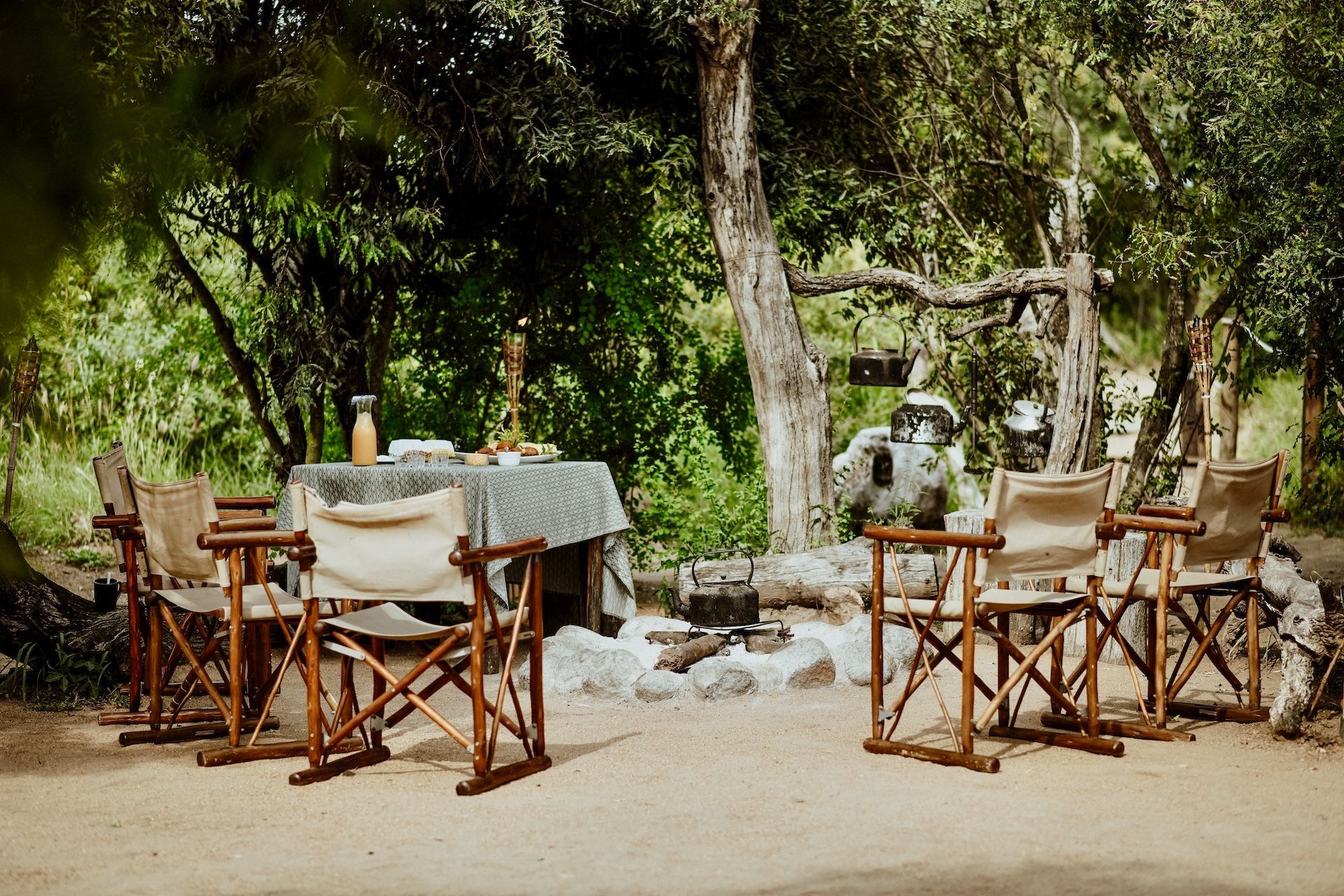A dining set with four canvas director chairs arranged around a small table in a wooded area with a central fire pit.