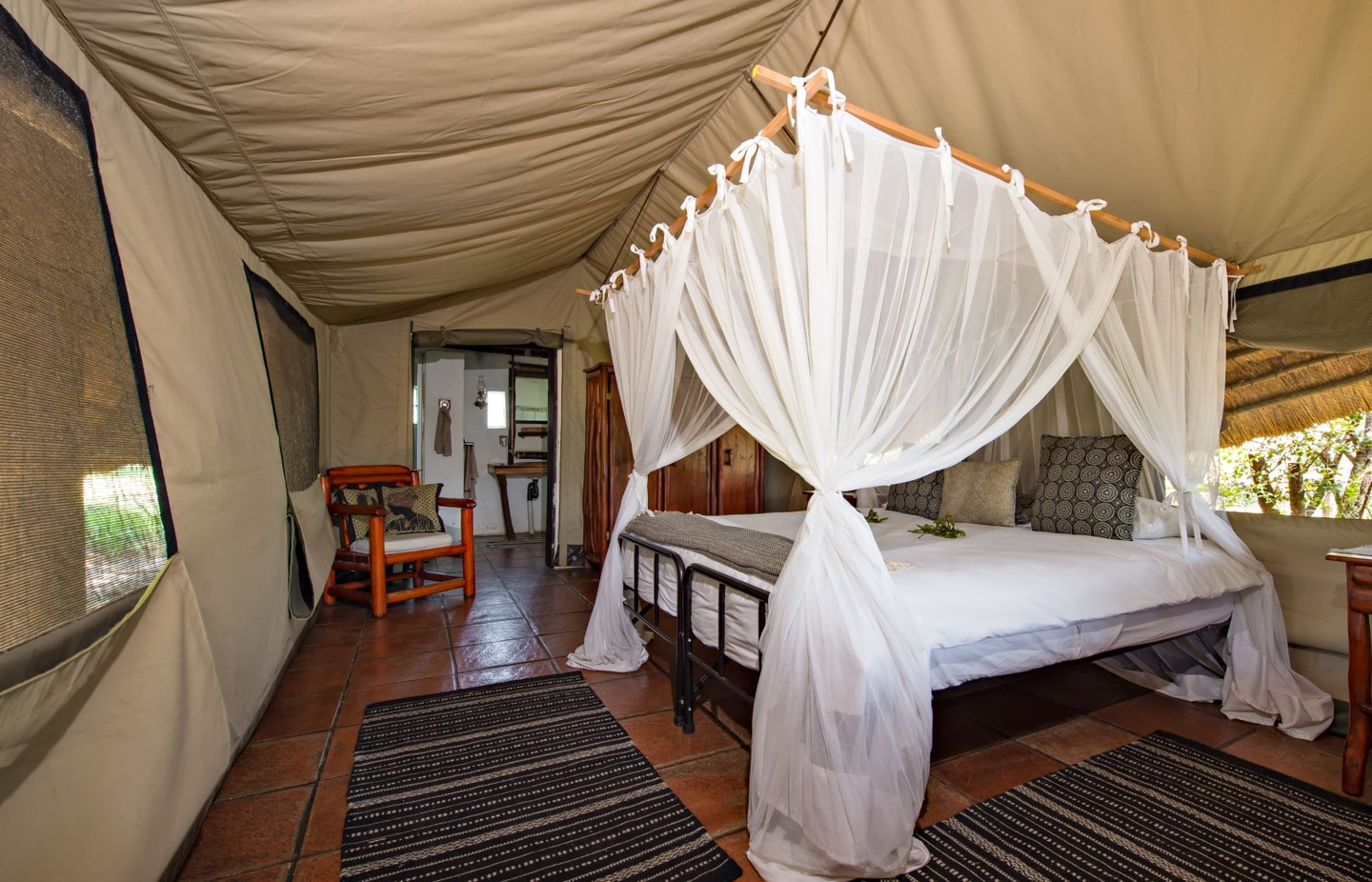 A luxury safari tent interior with a canopy bed, white linens, mosquito netting, wooden chair, and tiled floors.