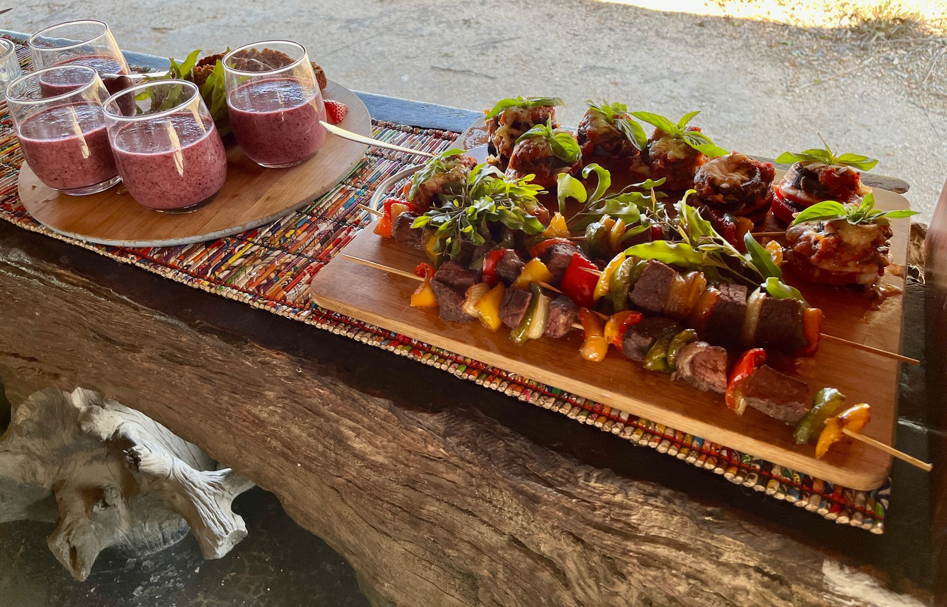 Skewers of meat and vegetables alongside glasses of purple smoothies arranged on wooden boards on a rustic surface.