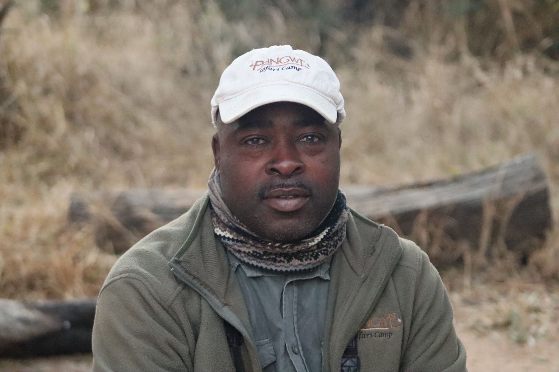 A person wearing a white baseball cap and olive fleece jacket with a neck gaiter, centered against a blurred outdoor scene.
