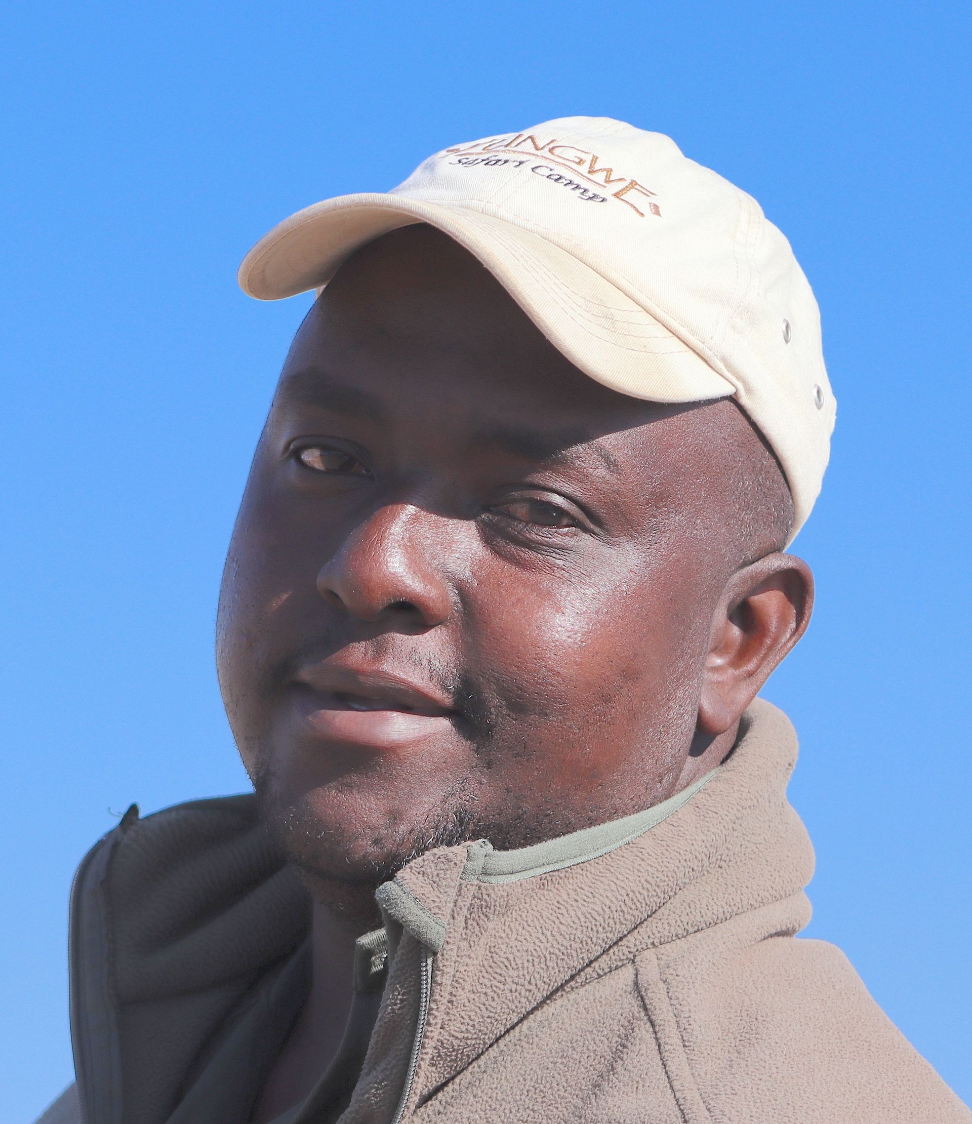 A person wearing a light-colored baseball cap and a brown fleece jacket, looking towards the camera against a blue sky.