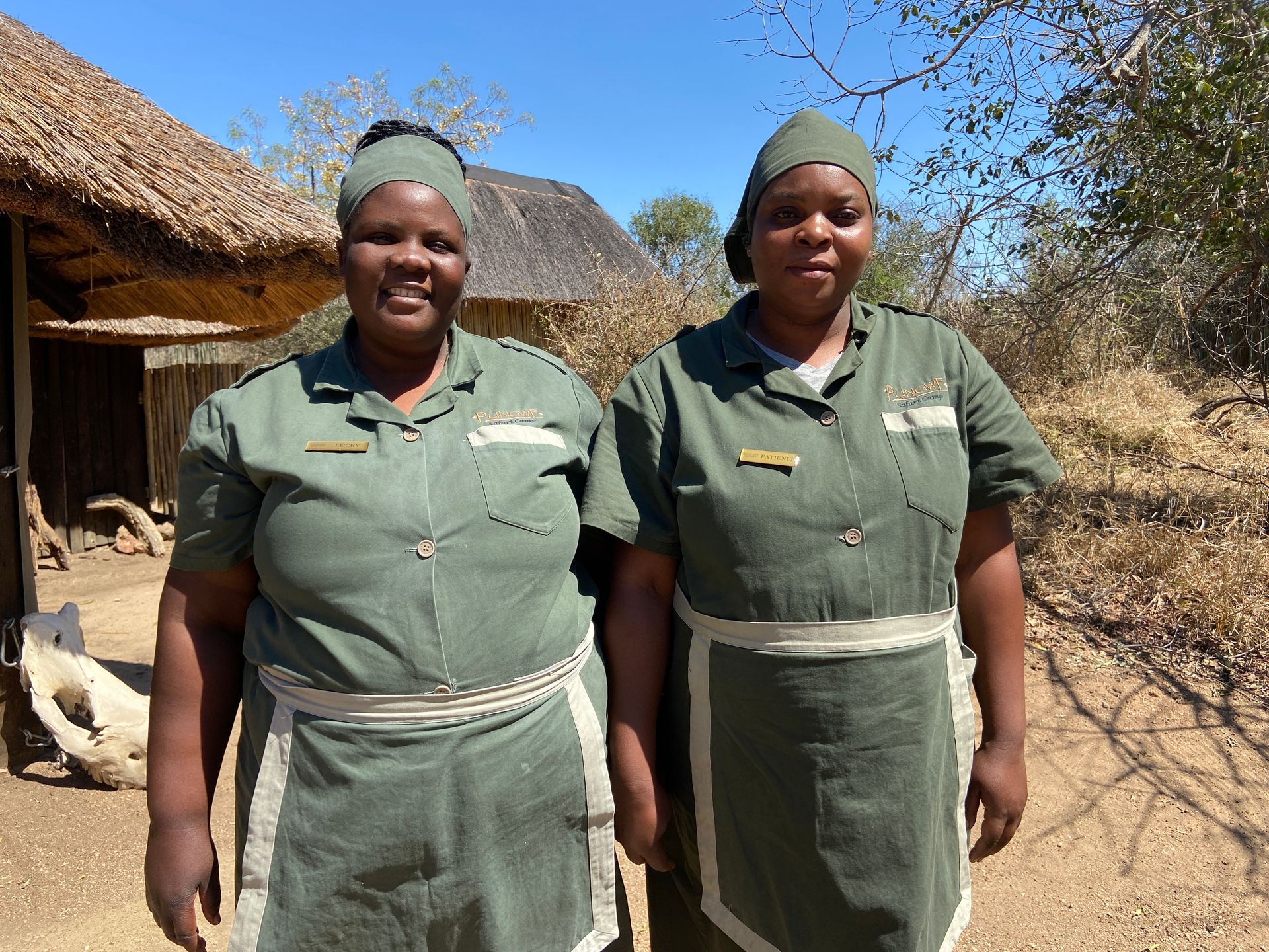 Two people in green uniforms and headscarves, standing outdoors in front of a rustic hut with a thatched roof.