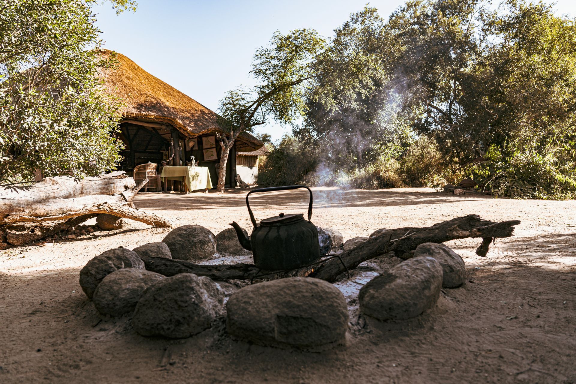 A black kettle sits inside a stone fire ring on sandy ground in front of a rustic hut with a thatched roof.