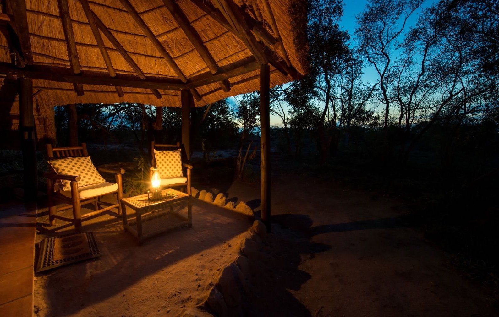 A dimly lit thatched patio with two chairs and a small table holding a lantern, overlooking trees at twilight.