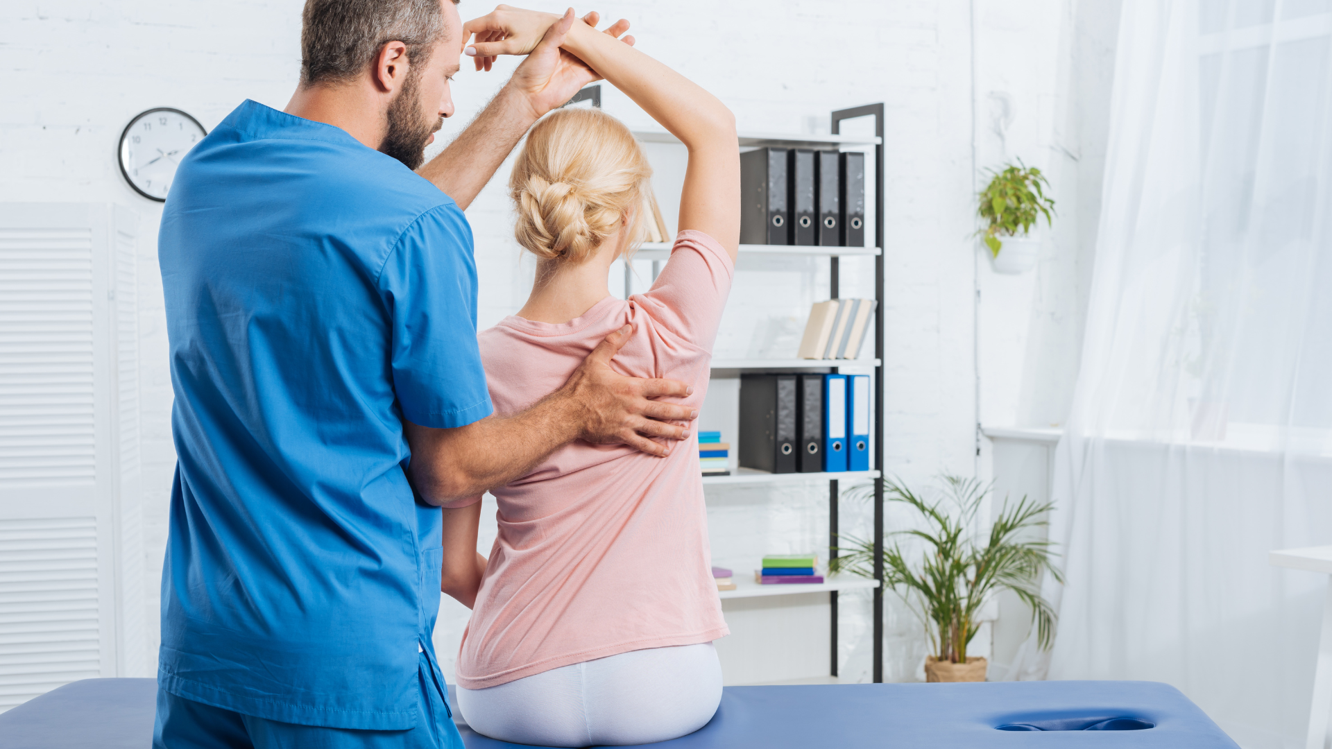Doctor in blue scrubs assisting a patient with arm movement in a medical office.