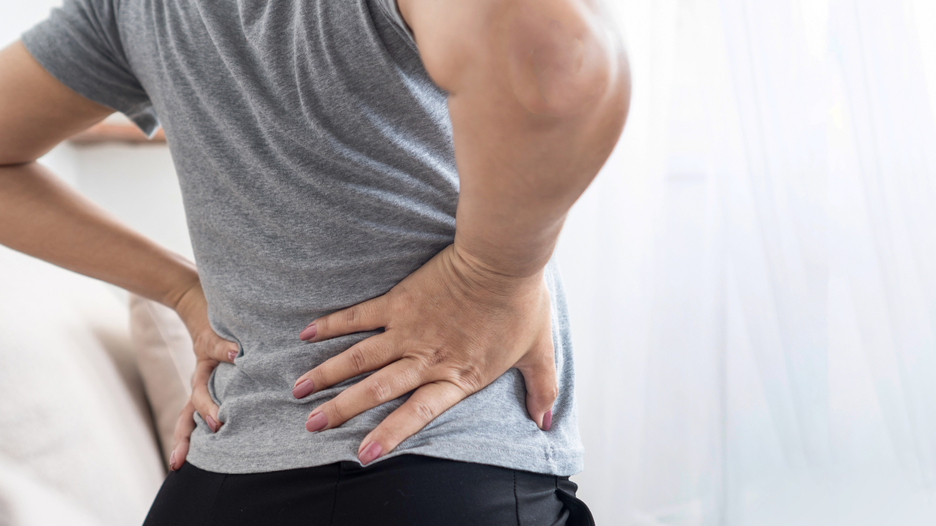 Person holding lower back in pain, standing indoors on a bright background.