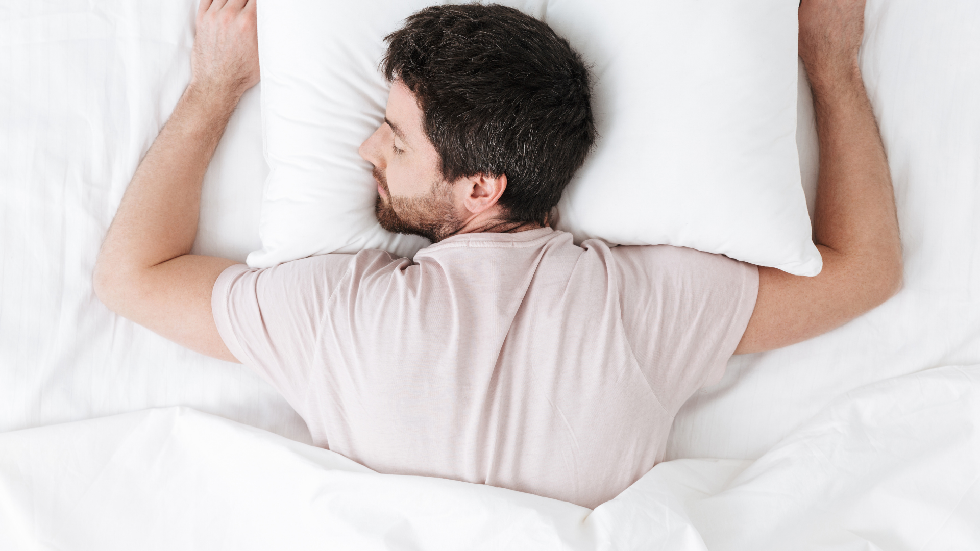 Man lying on a white bed on his stomach, arms raised, looking to the side