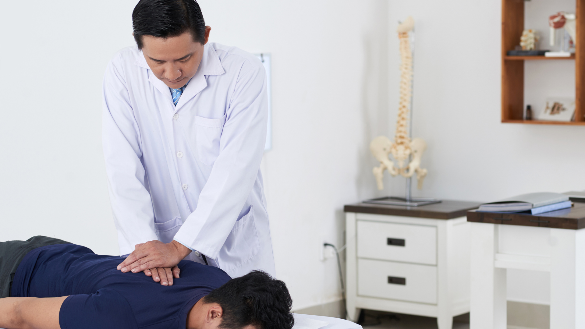 A doctor in a white coat examines a patient's back on a table in a clinic, with a spine model in the background.