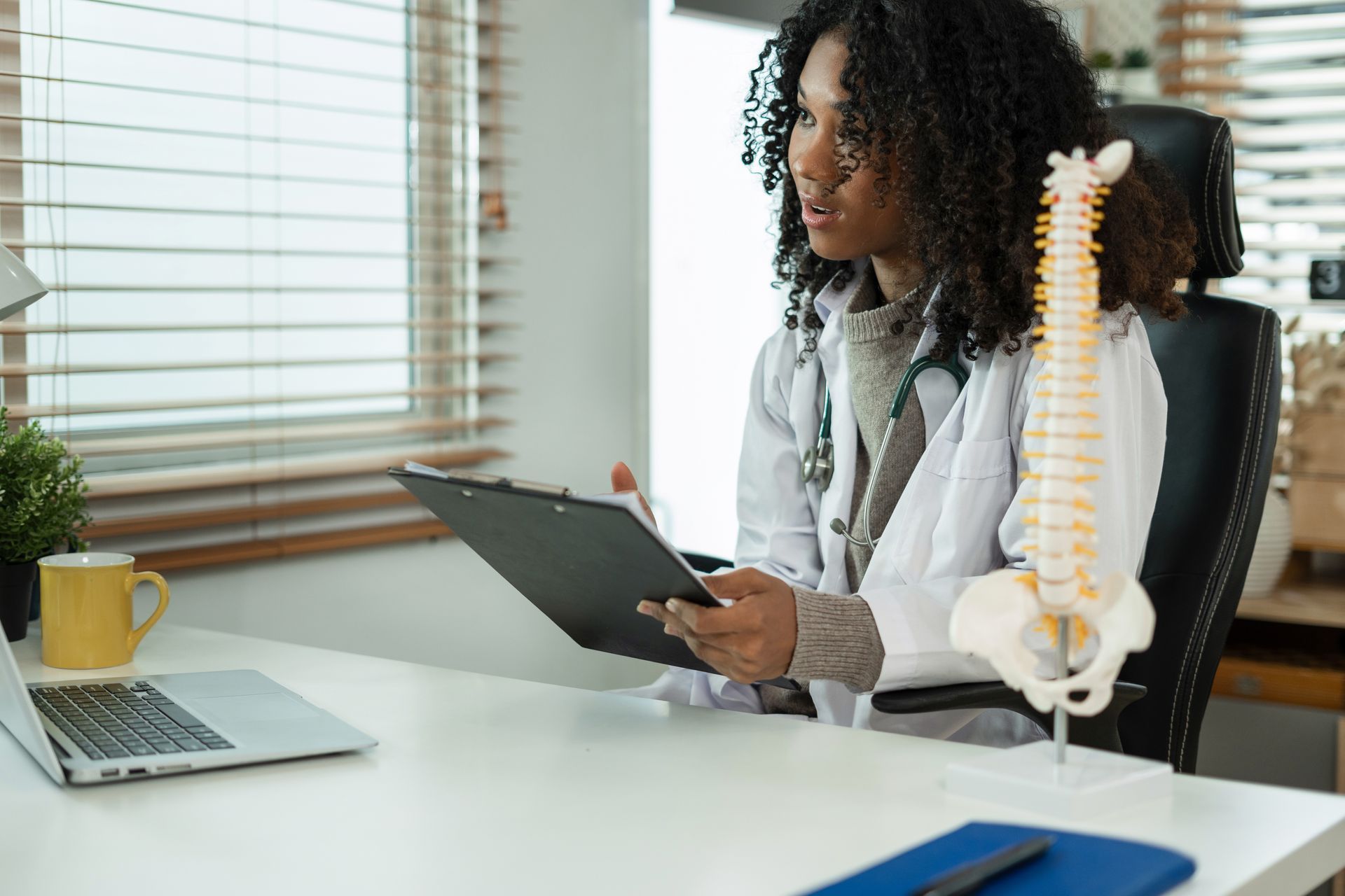 A doctor is sitting at a desk with a clipboard and a model of a spine.