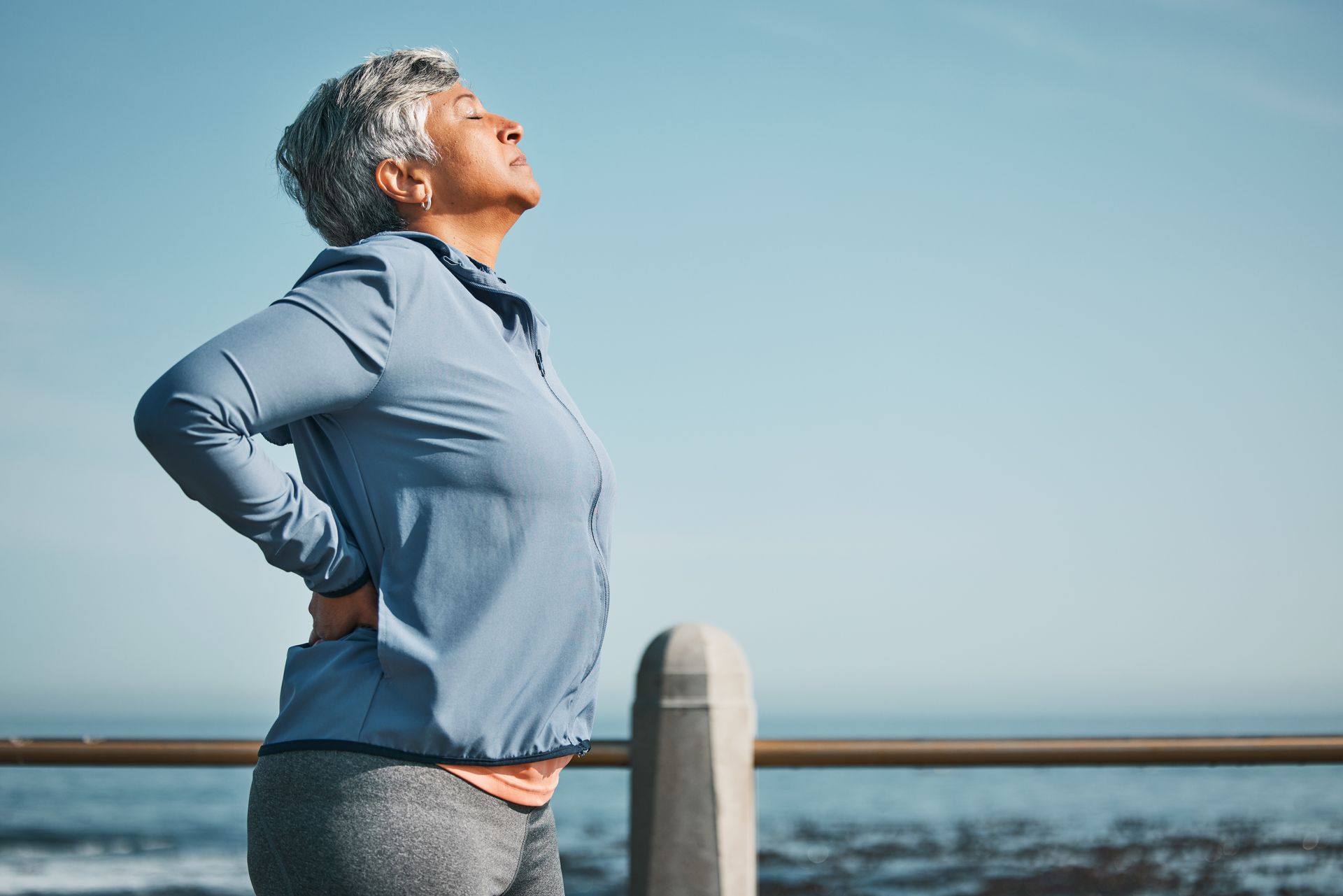 An older woman is standing on a pier holding her back in pain.
