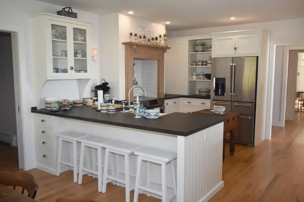 A kitchen with white cabinets , stools , a refrigerator and a sink.