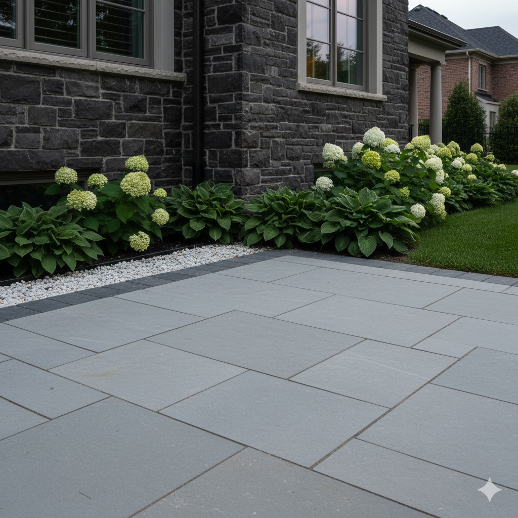 Gray stone patio with black trim, adjacent to a stone-clad building and green shrubbery with white flowers.