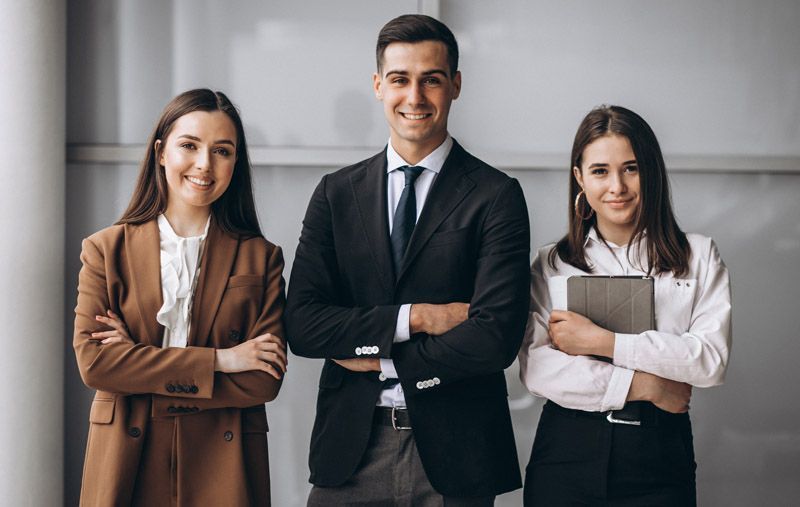 Three professionals standing with arms crossed in a bright office, wearing business attire.