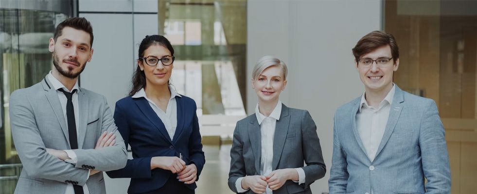 Four business professionals stand together in a modern office, posing with arms crossed and smiling.