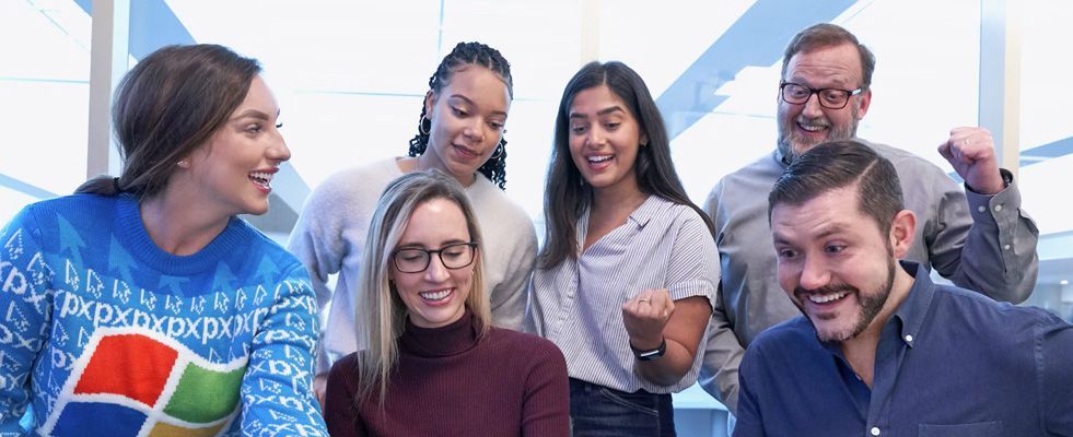 Six coworkers smiling around a laptop, with one wearing a blue sweater and another giving a thumbs-up