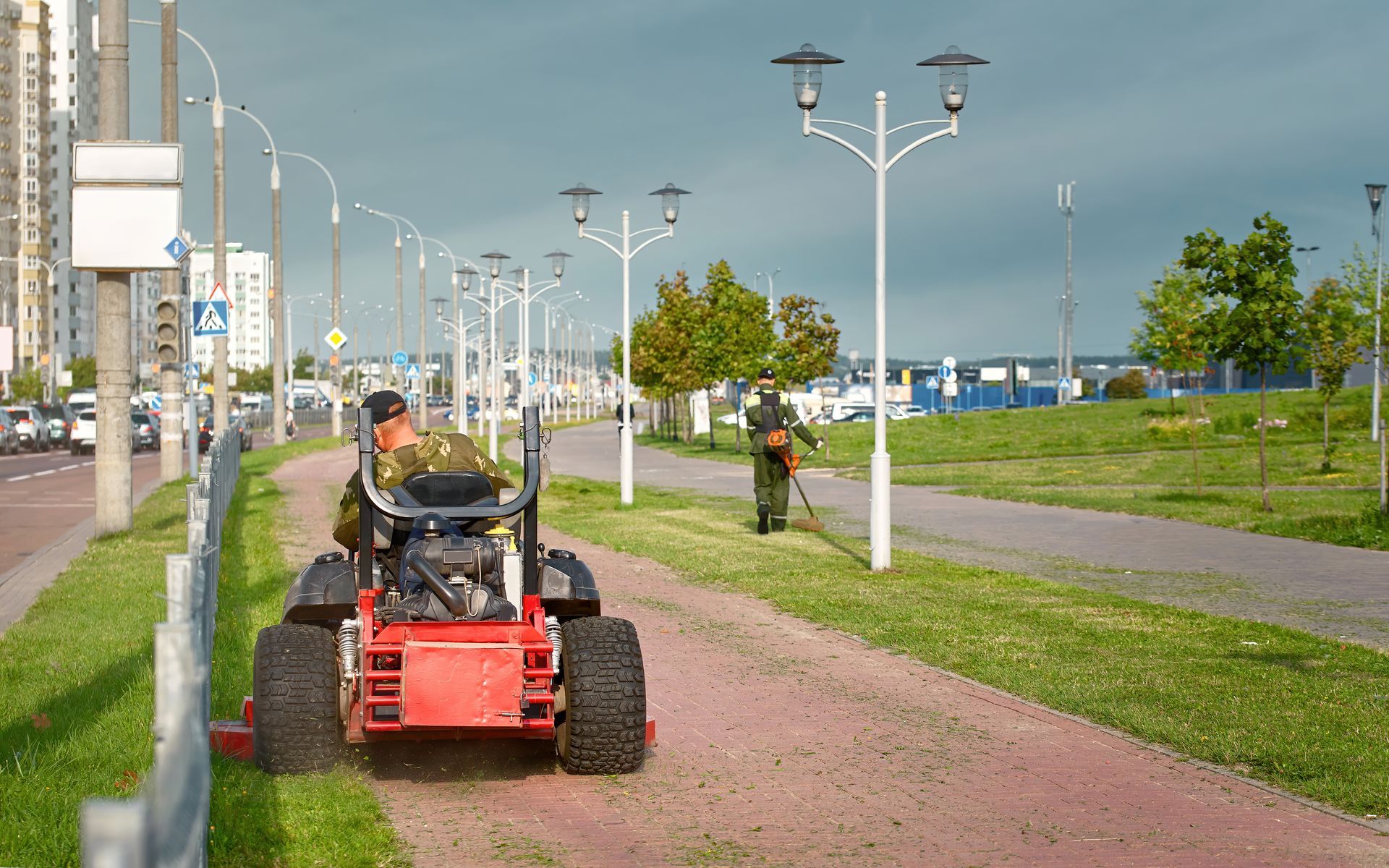 Person on a red utility vehicle on a paved path beside a grassy waterfront promenade with streetlights
