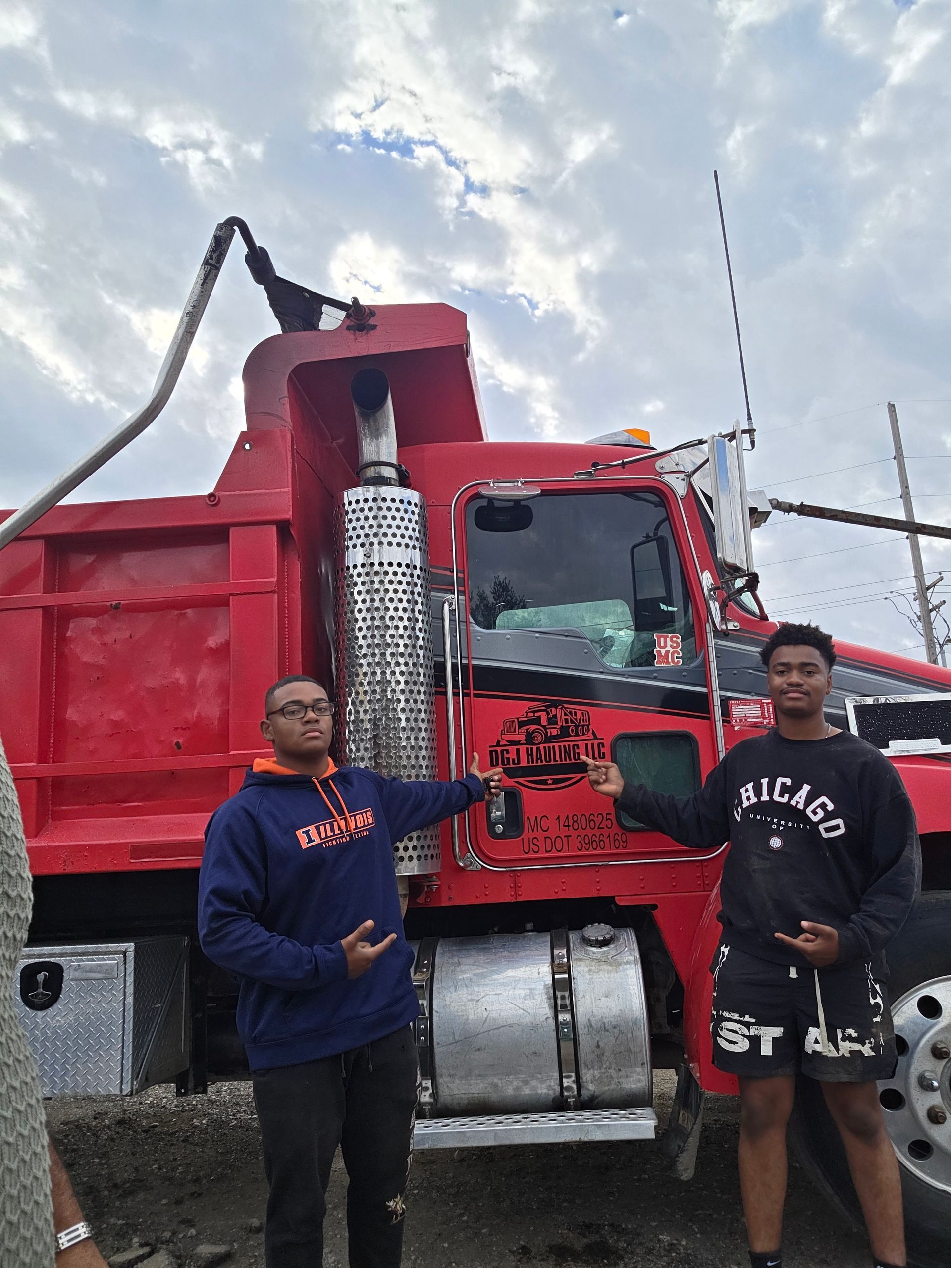 Two men stand beside a red dump truck, pointing. Cloudy sky in the background.