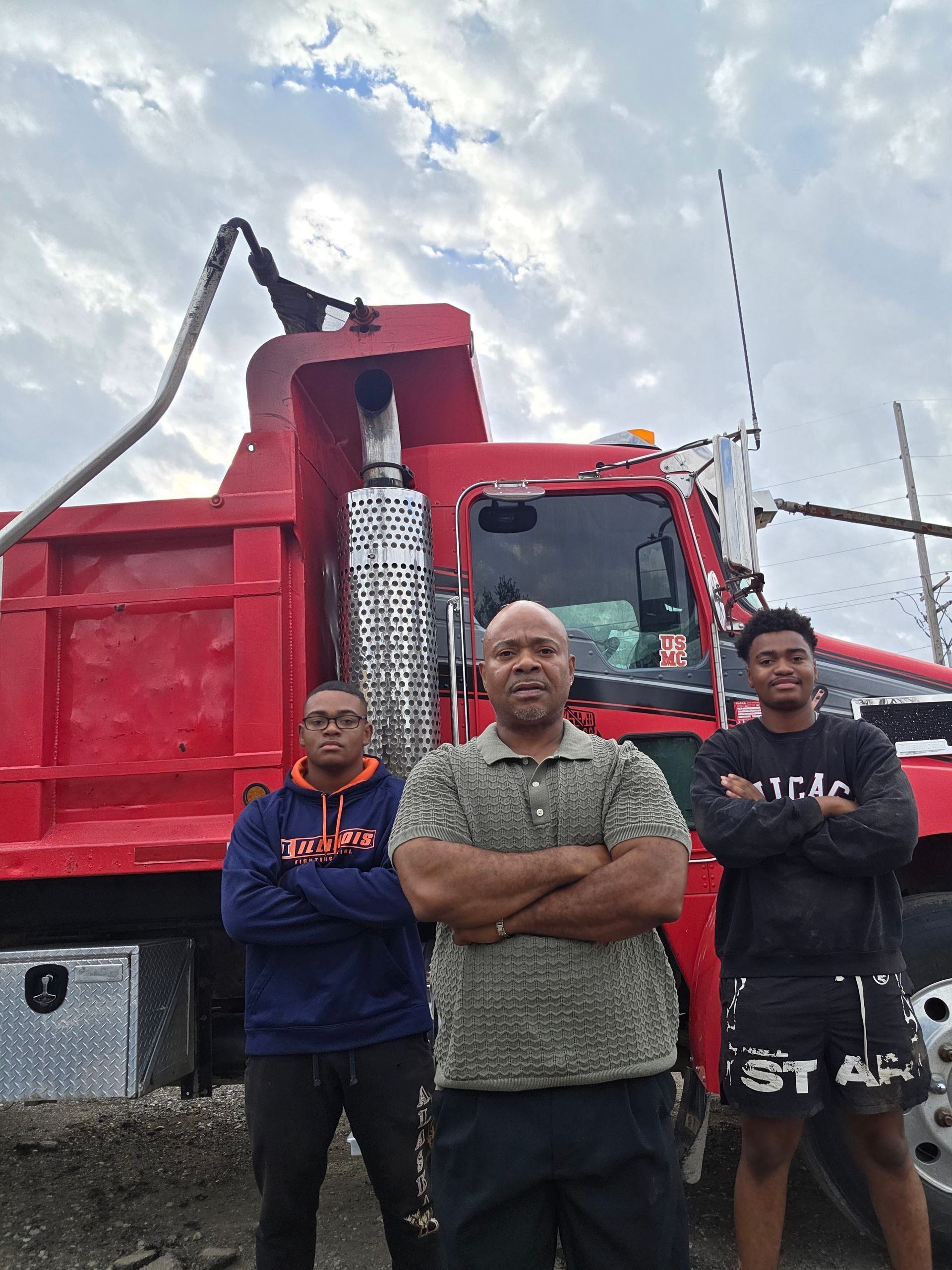 Three men stand in front of a red dump truck, arms crossed. Cloudy sky, industrial setting.