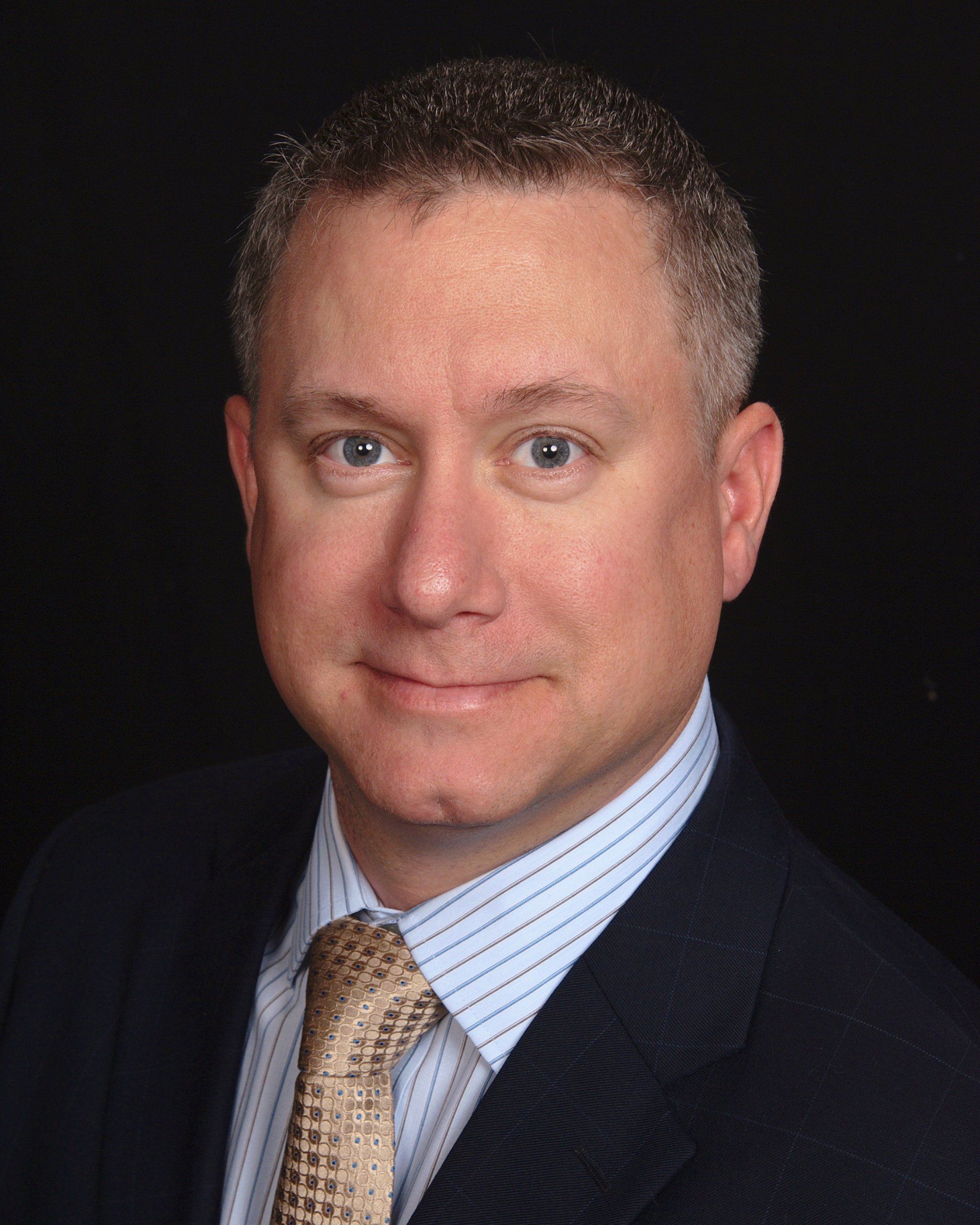 Studio headshot of a man in a dark suit and tie against a black background