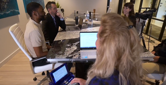 A white woman and a black man sit together at a table in an office setting and look over documents together.