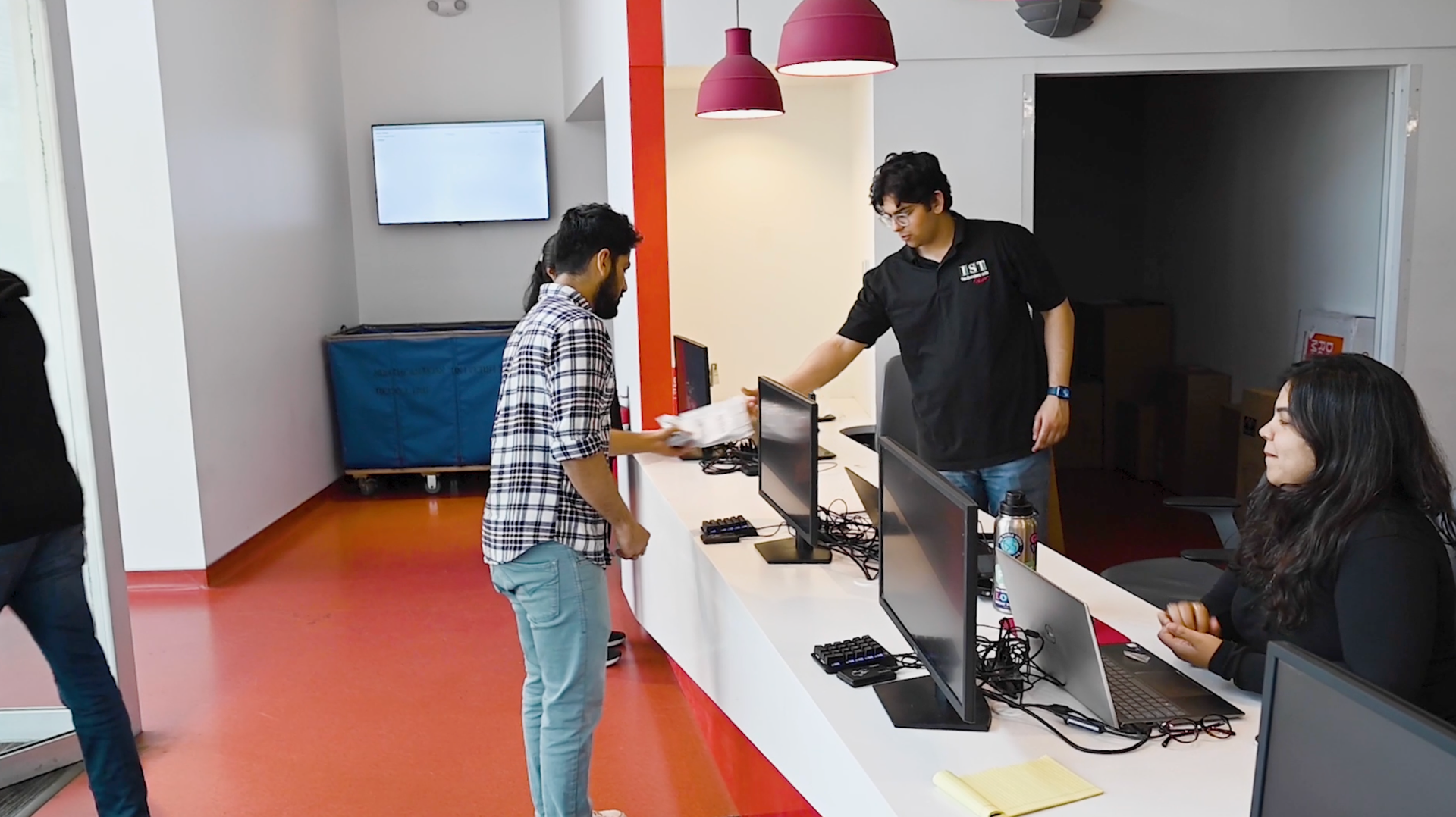 People at a computer check-in desk in a bright office with red floor and hanging pink lamps