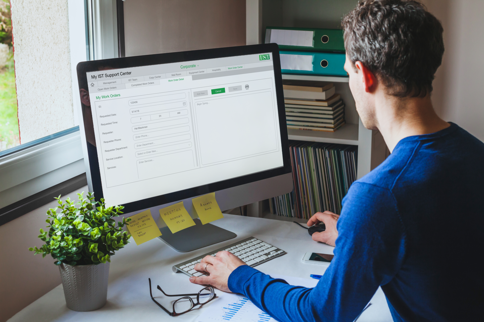Person working at a desk with a large monitor, keyboard, sticky notes, and a small potted plant