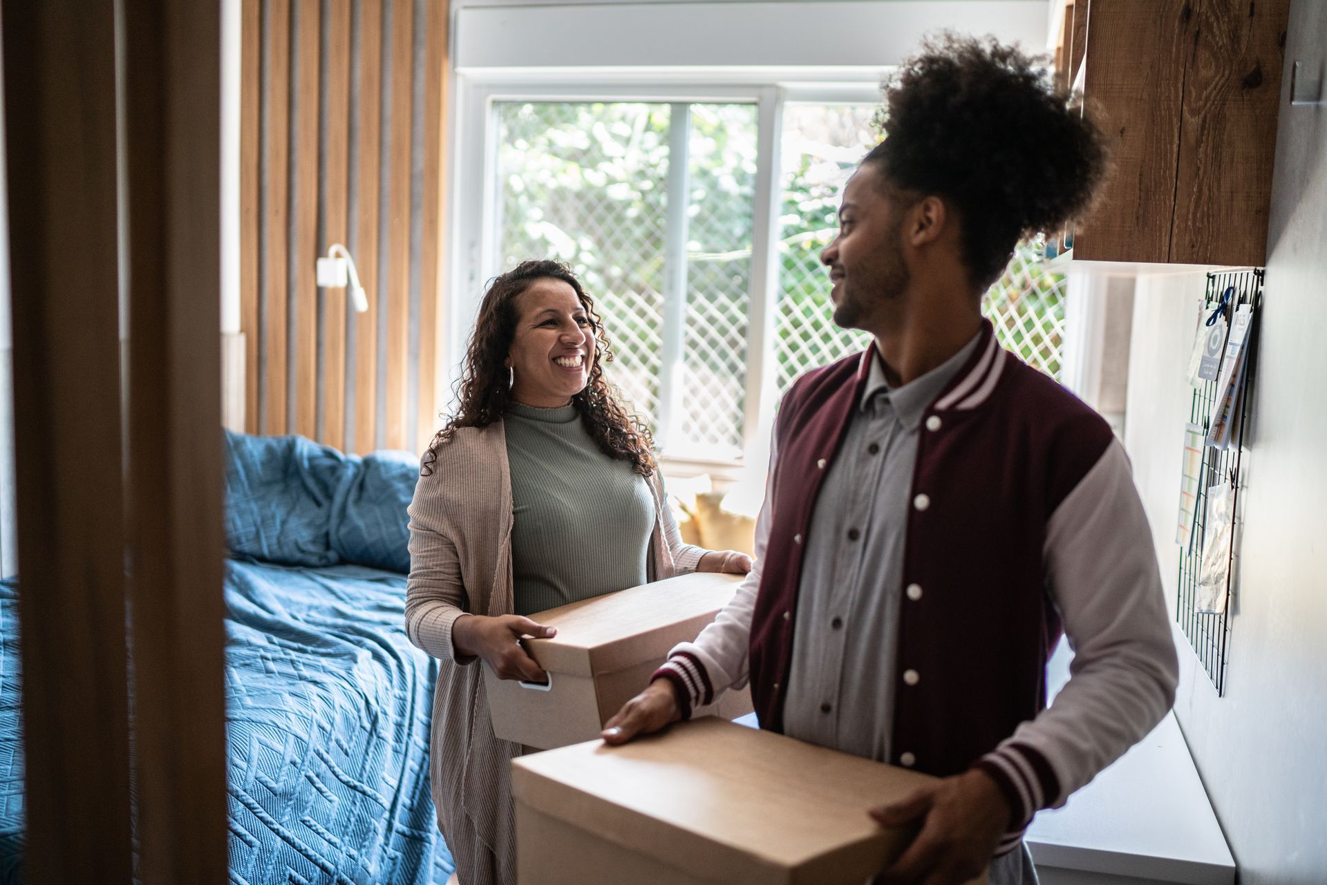 Two people carrying boxes in a bright bedroom with a blue bed and window light