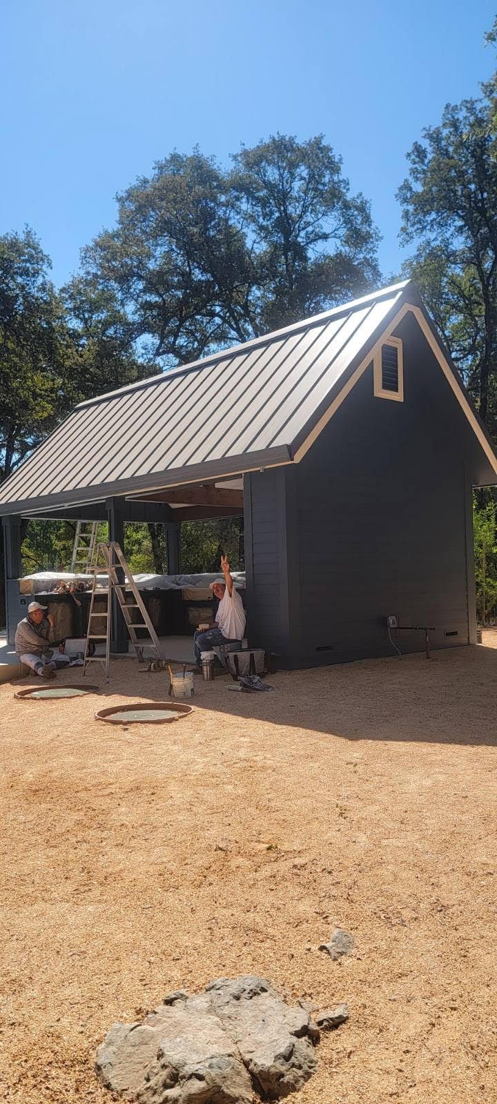 Small dark shed with metal roof and porch in a sunny gravel yard with trees in the background