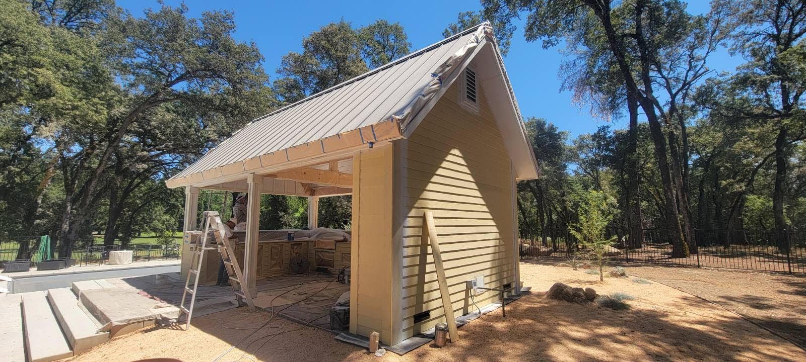 Small yellow shelter with a metal roof in a wooded outdoor area, set on sandy ground.