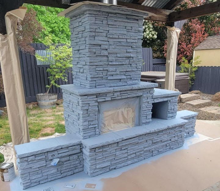 Stone outdoor fireplace and grill structure in a backyard, with a tall chimney and built-in shelf.