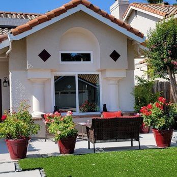 Front view of a house patio with red potted flowers, a bench, and green lawn.