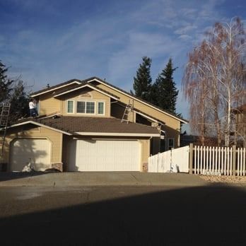 Suburban house with beige siding, white garage doors, and a white fence under a blue sky.
