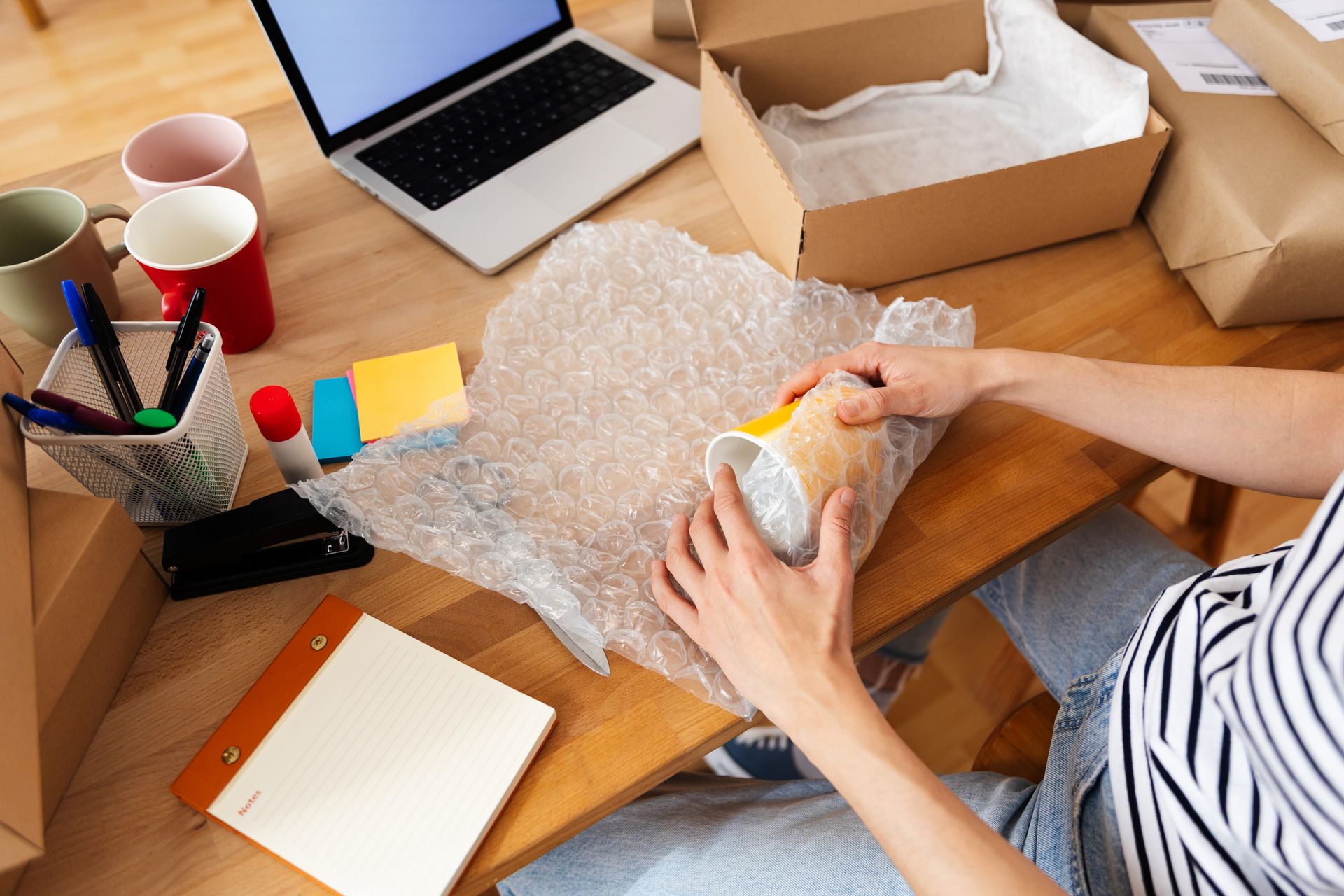 Person wrapping a mug in bubble wrap at a desk with boxes and a laptop. Person wrapping a mug in bubble wrap at a desk with boxes and a laptop.